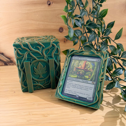 Green decorative box with a card holder containing a trading card on a wooden surface with a plant in the background.